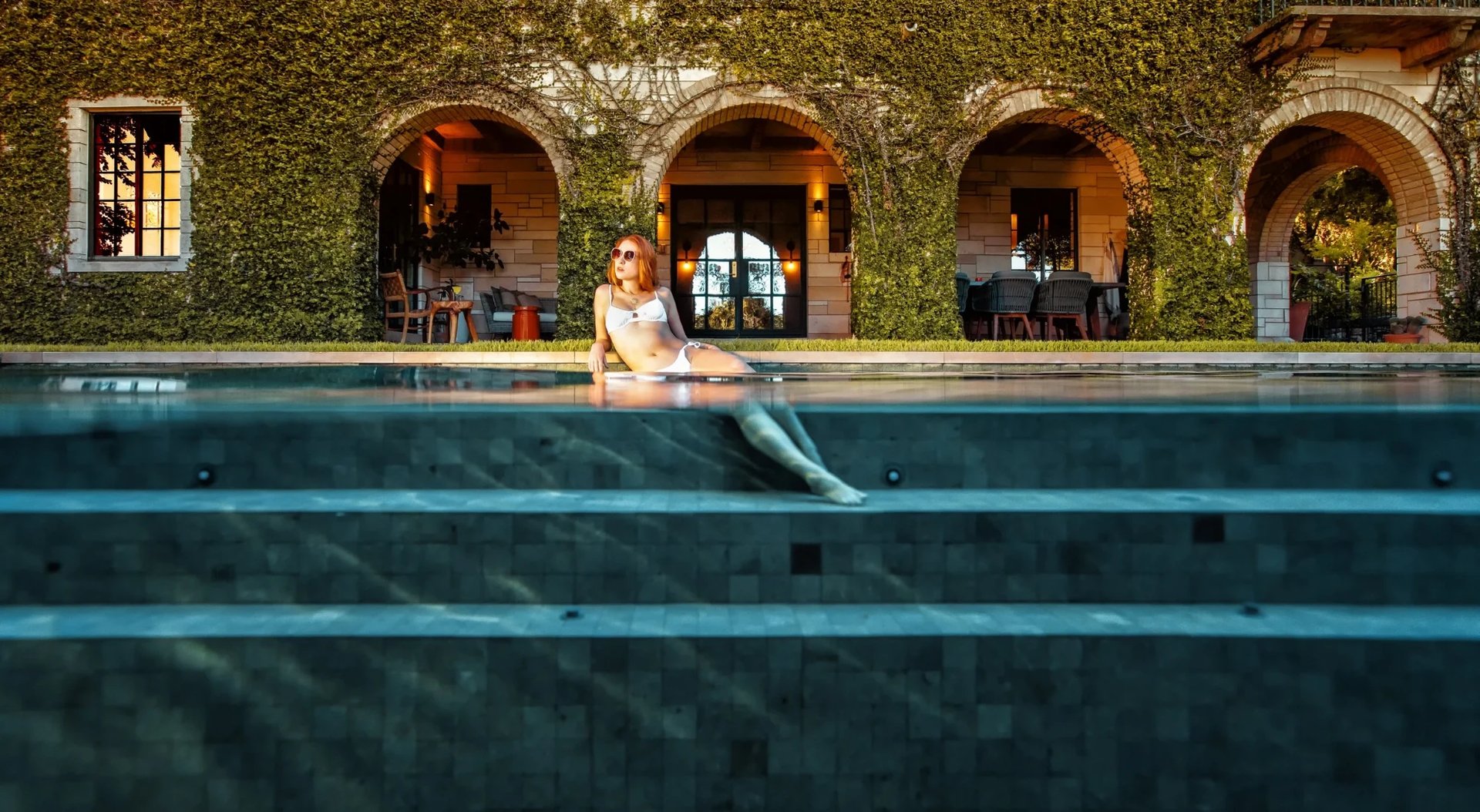 Woman in a white suit reclining on the steps of a tiled lap pool before an ivy-draped arched loggia