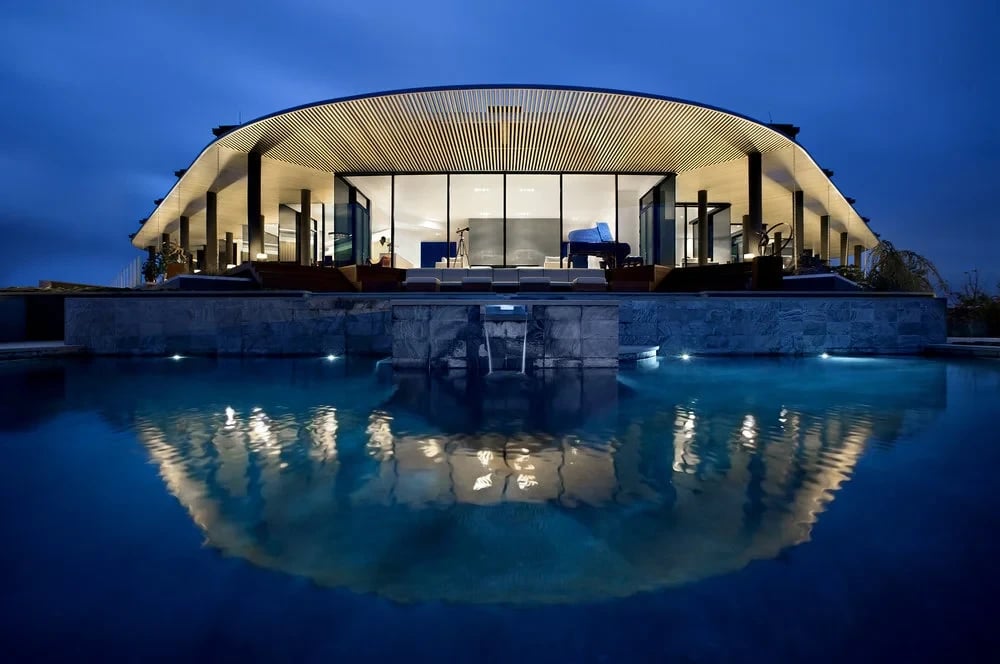 Curved-roof glass home glowing at twilight above a stone-walled pool with a spillway, reflected in still water