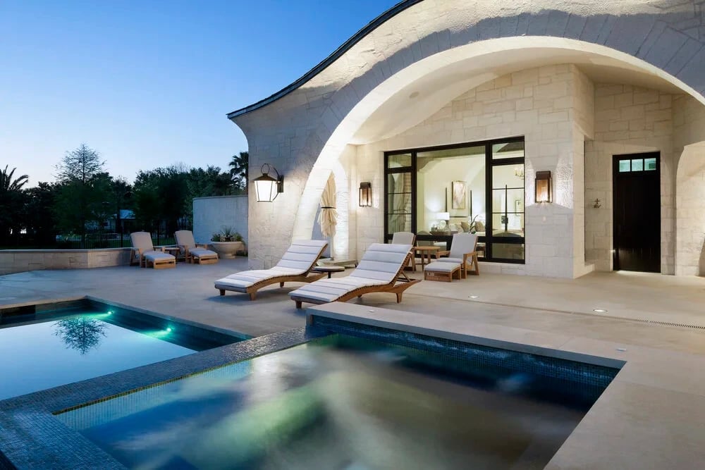 Geometric pool and raised spa with mosaic tile beside lounge chairs under an arched white-stone loggia