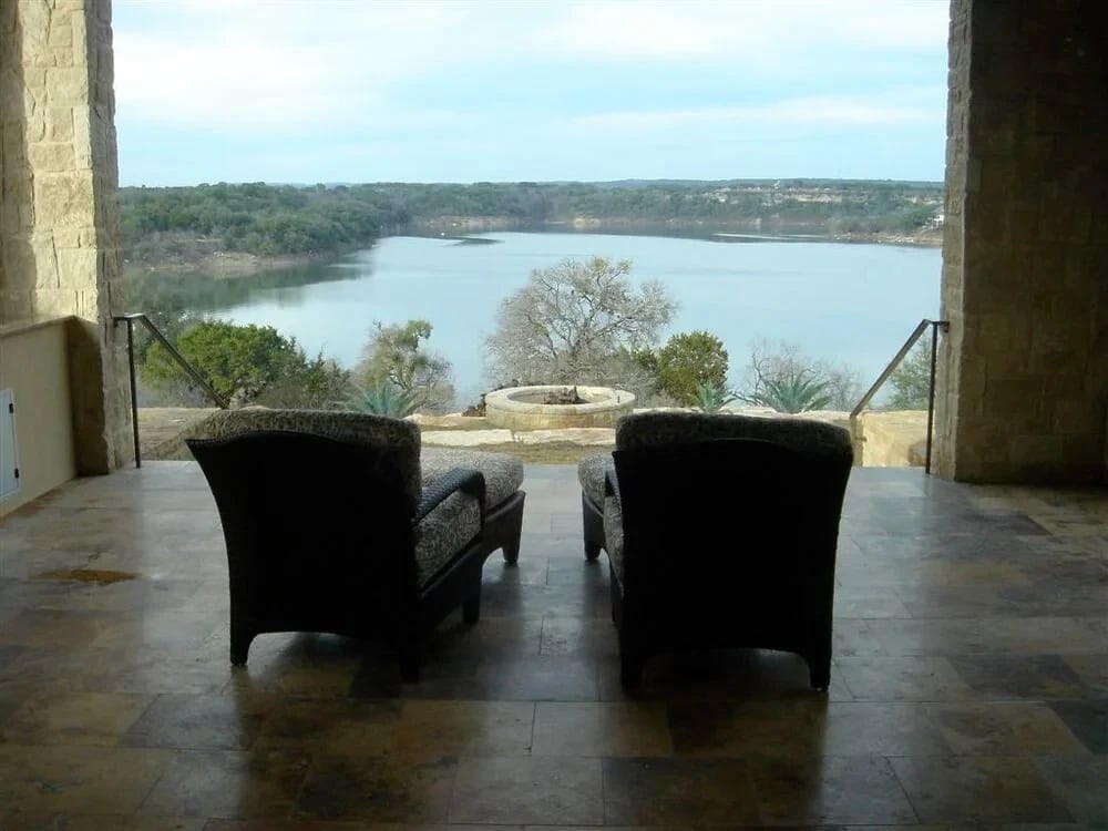 Two wicker chairs on a travertine patio framing a wide lake and wooded hills under stone arches
