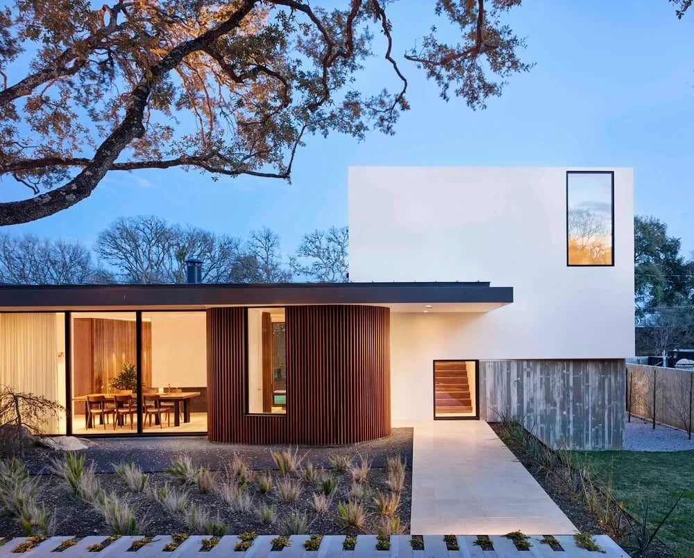 Crisp modern home with white and wood walls and a curved glass corner under oaks at dusk
