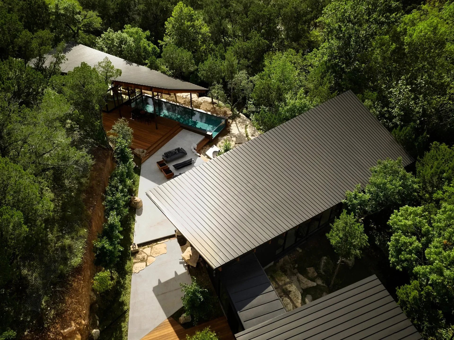 Aerial view of a hillside home with metal roofs and a glass-walled pool pavilion tucked into thick green woods