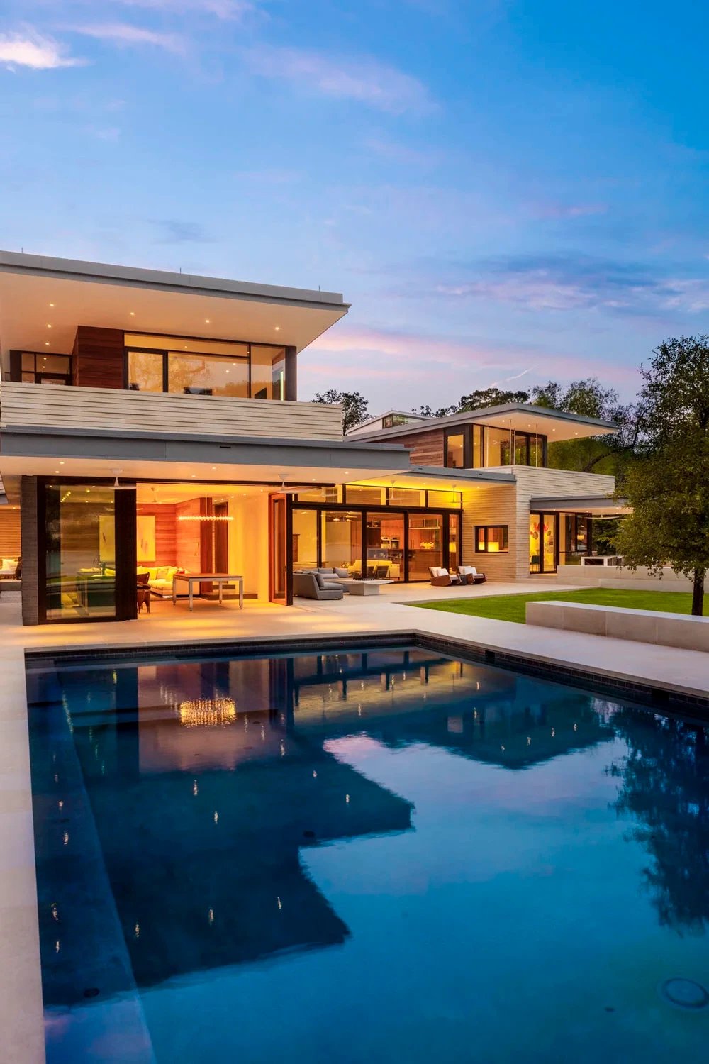 Geometric pool reflecting a lit two-story modern home with glass walls at dusk