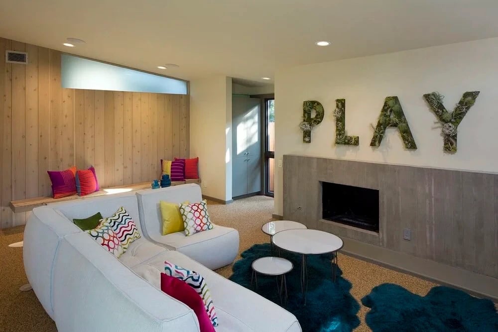 Midcentury living room with cedar paneling, white sofas, a concrete fireplace and a teal rug spelling PLAY
