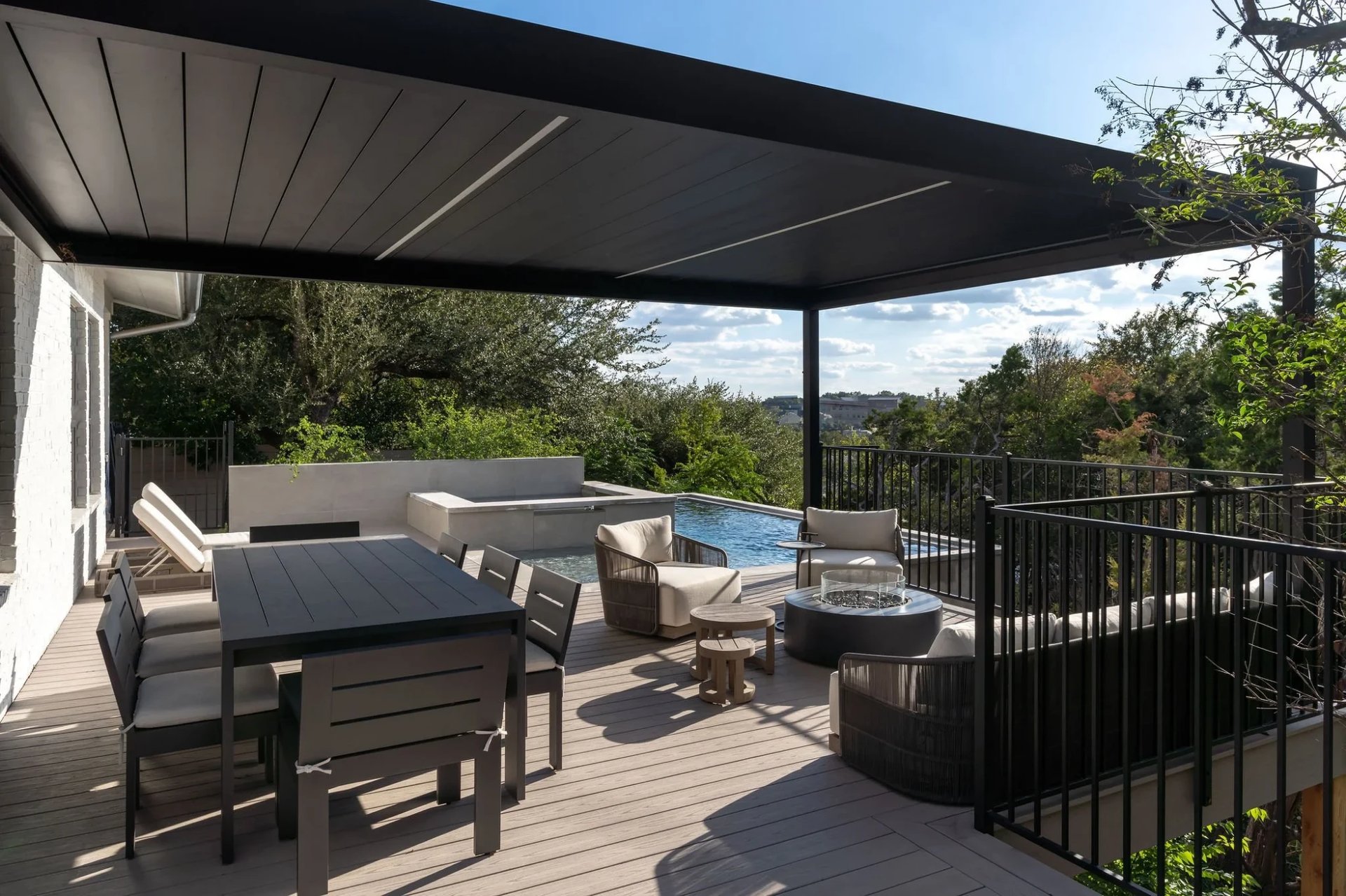 Covered deck with a black pergola, dining set, and fire pit beside an infinity pool over the trees