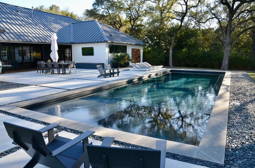 Geometric pool with dark interior and stone coping beside a metal-roof home and patio