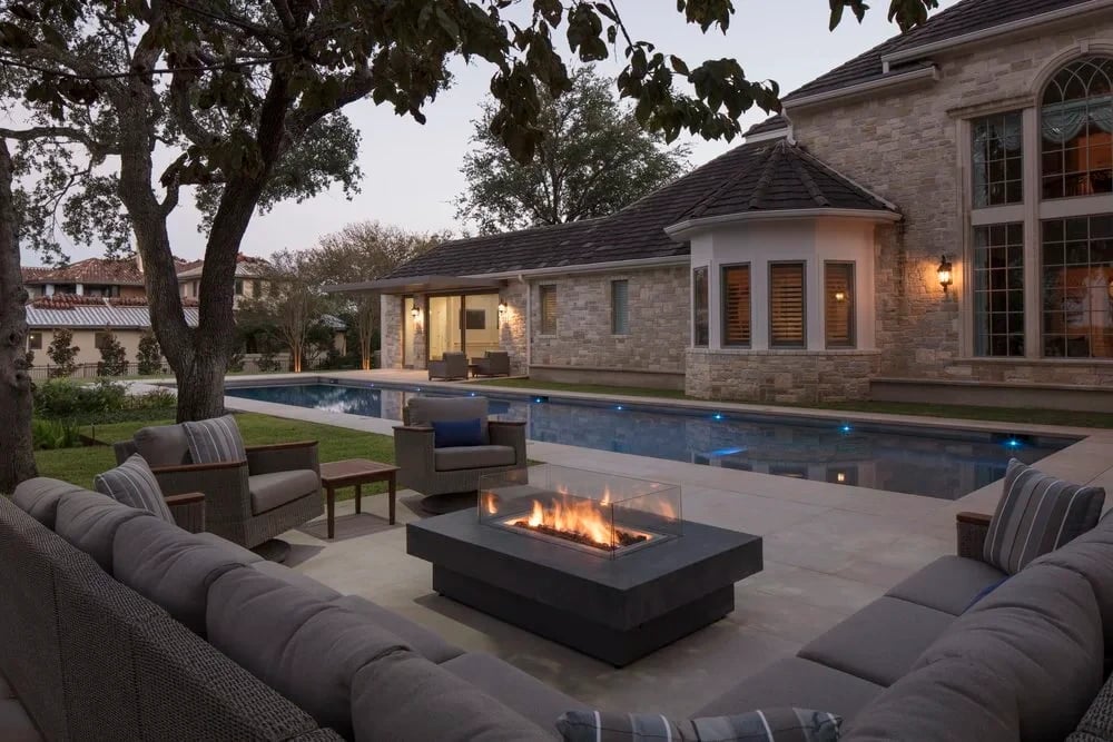 Patio sectional around a linear gas fire table at dusk, with a geometric pool and stone home behind