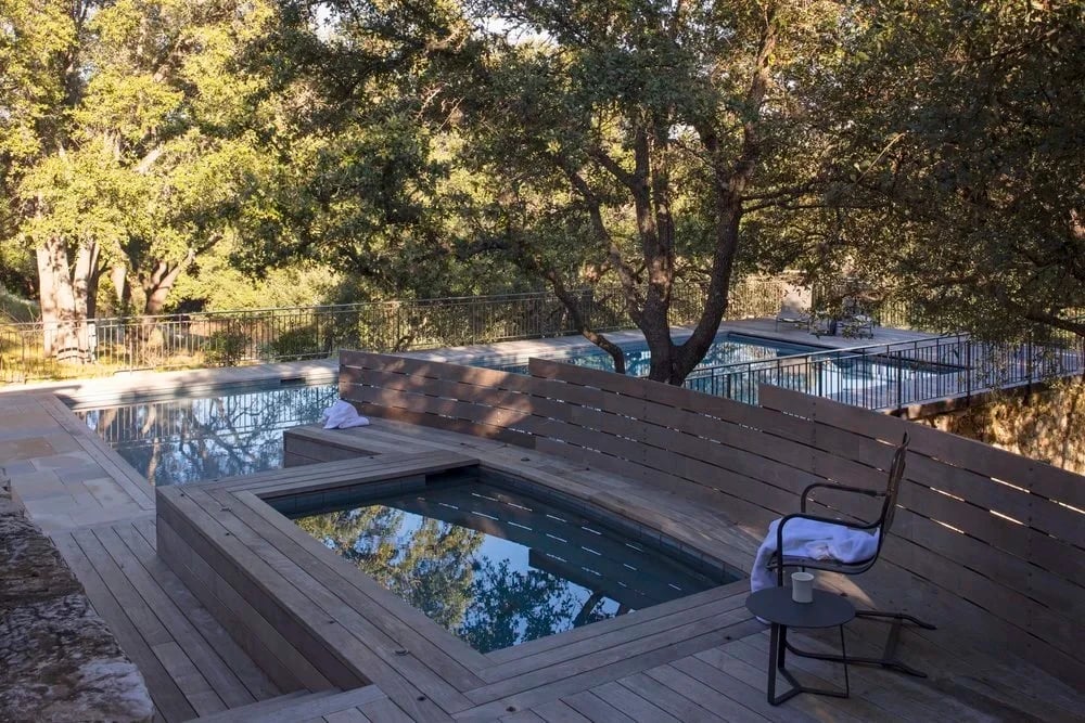 Wood deck wrapping a geometric pool and spa among live oaks on a sloped Hill Country lot