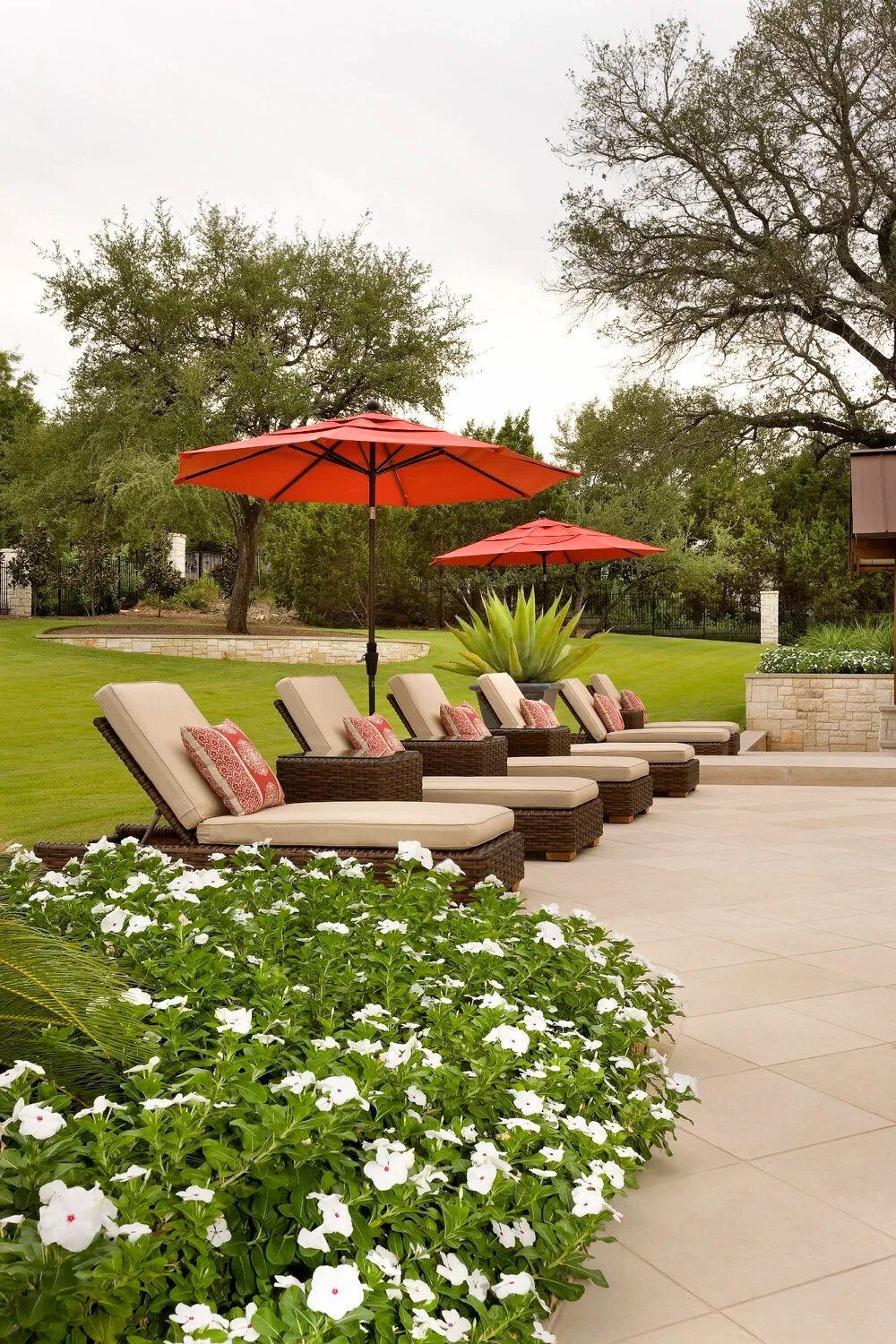 Row of cushioned chaise lounges under red umbrellas on a limestone patio beside flowering vinca