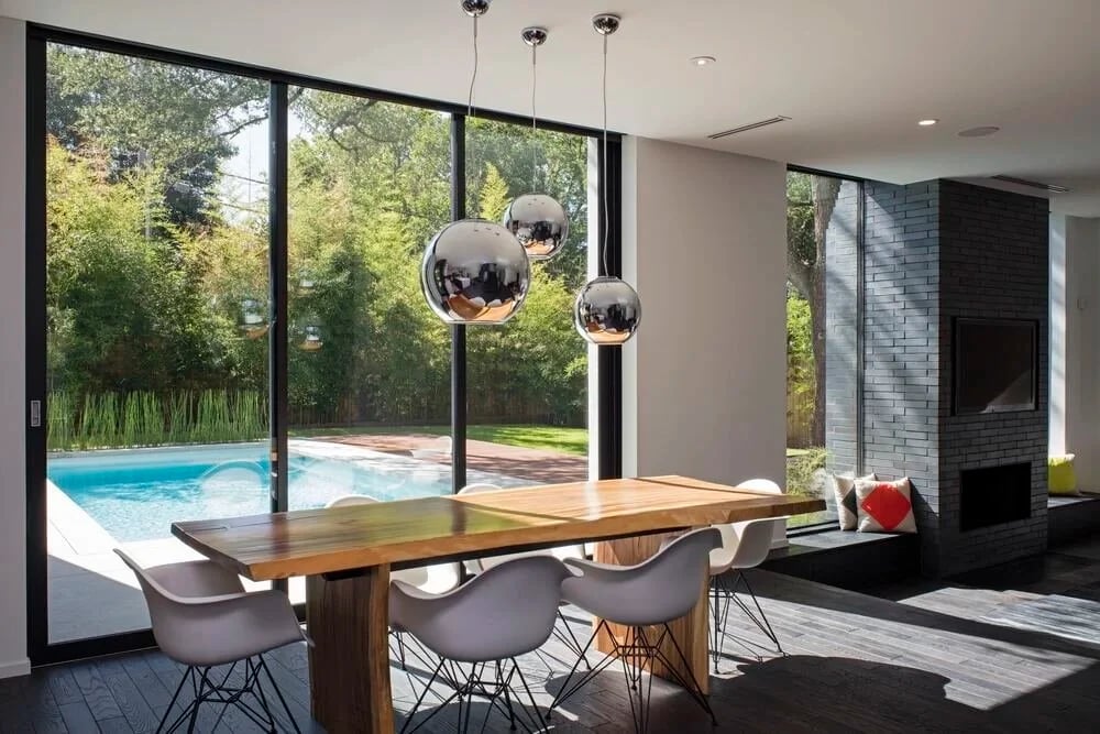 Modern dining room with a live-edge wood table, chrome globe pendants, and floor-to-ceiling glass facing the pool