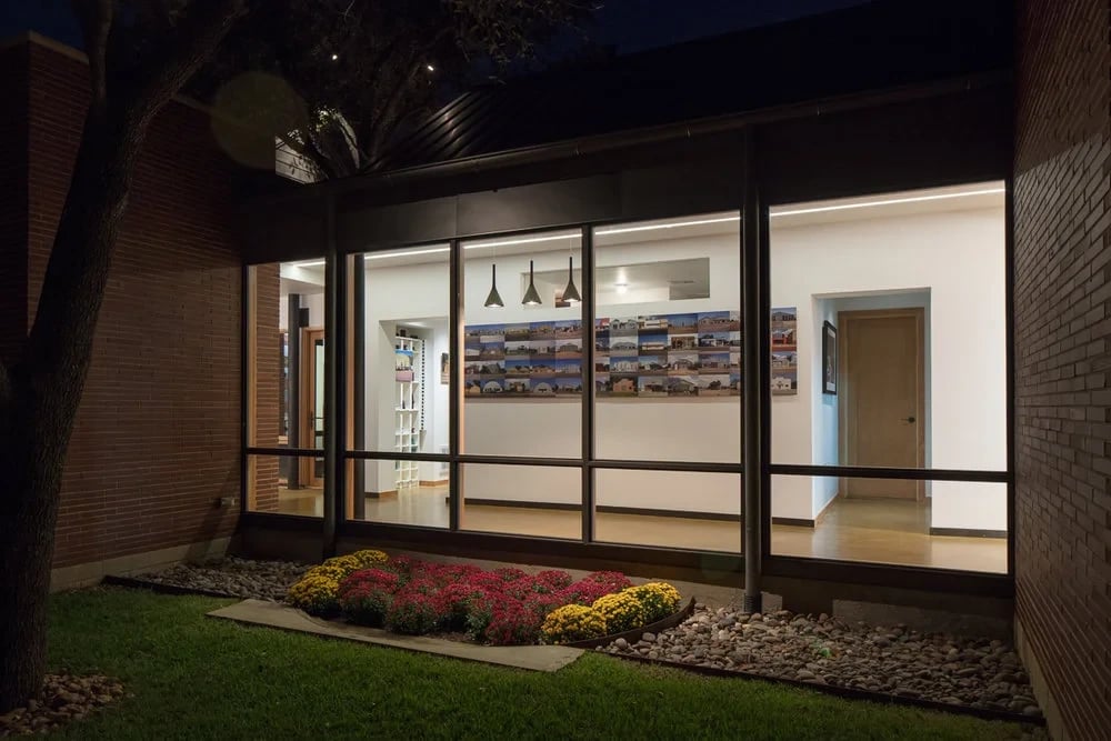 Lit glass-walled room at night seen from a brick courtyard, with red and yellow blooms and a tree