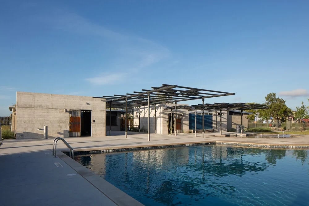 Concrete bathhouse with steel pergolas at dusk, mirrored in a still public pool