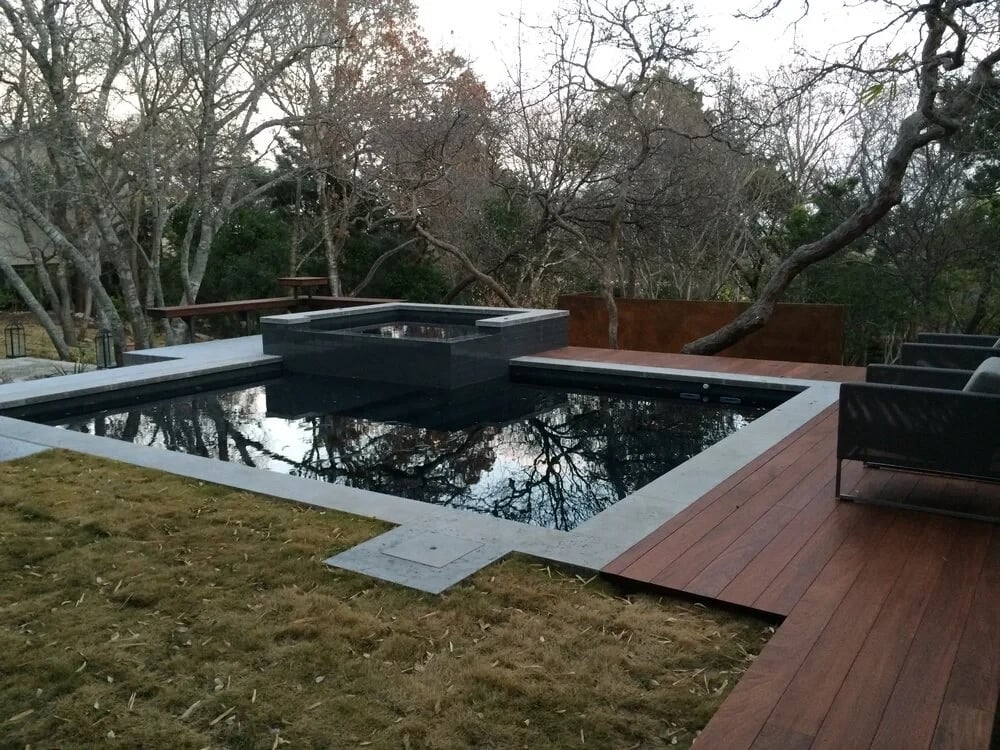 Dark geometric pool with a low spillway and wood deck reflecting bare winter trees in the backyard