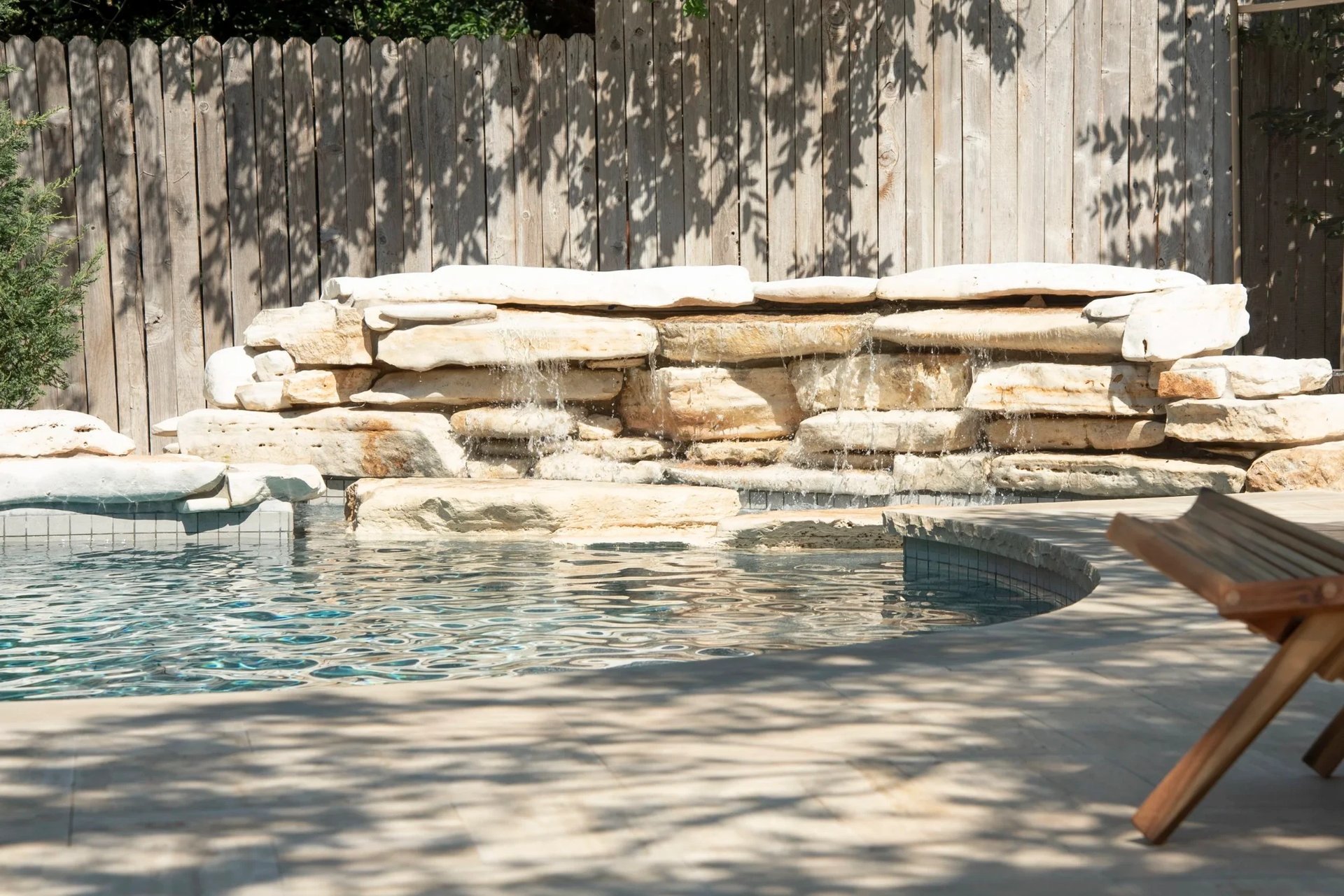 Stacked-stone grotto waterfall trickling into a freeform pool with a rounded spa and dappled shade