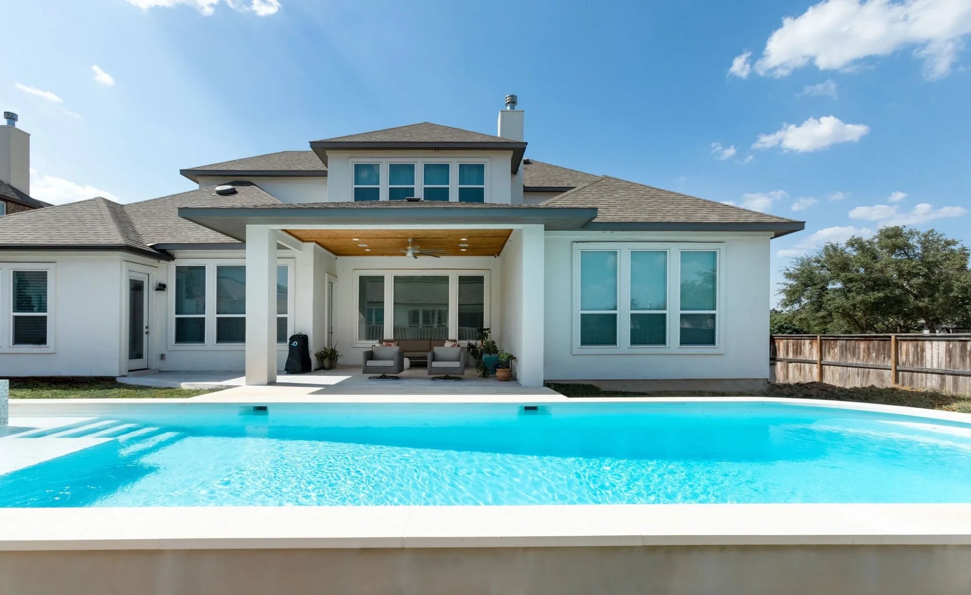 Geometric pool behind a two-story white stucco home with a covered patio and tall windows under a clear blue sky