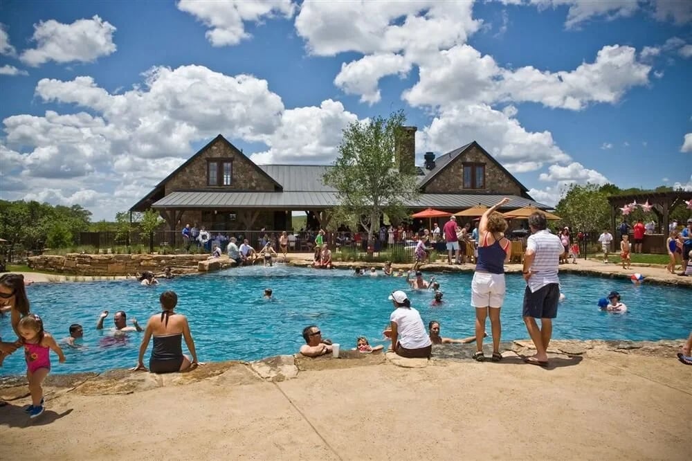 Crowd swimming and gathered around a large freeform pool at a stone lodge under puffy clouds