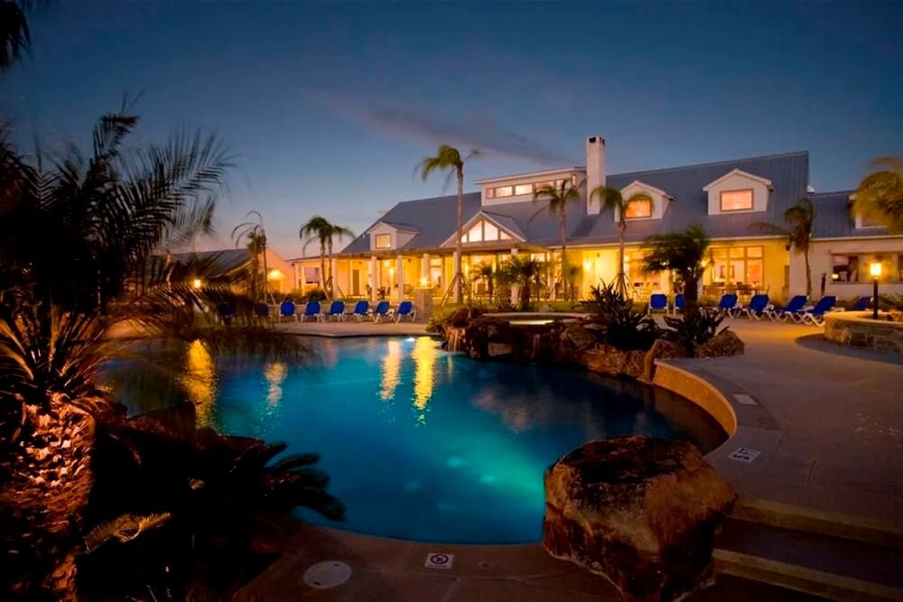 Freeform pool with a rock waterfall and grotto lit blue at dusk, palms and a lit lakeside lodge beyond