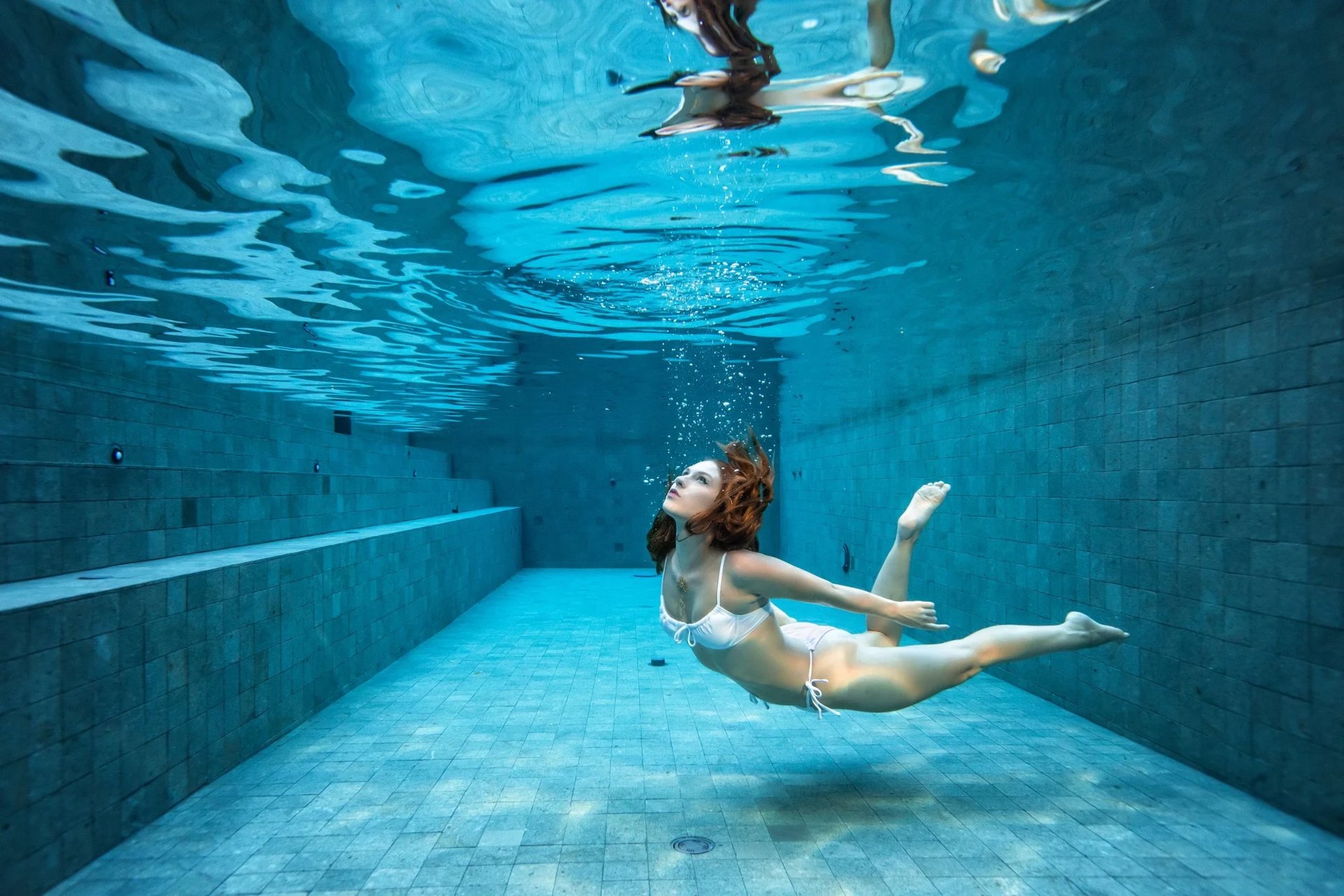 Woman lounging on the submerged steps of a green-tiled lap pool with an ivy-covered arcade behind