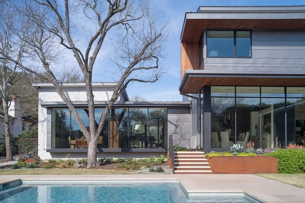 Modern home with steel siding and glass walls beside a bare tree, with a geometric pool edge in the foreground