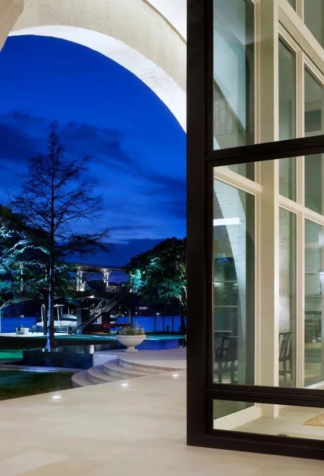 View through an arched doorway at dusk to a lakeside lawn, lit trees and a dock beyond glass doors