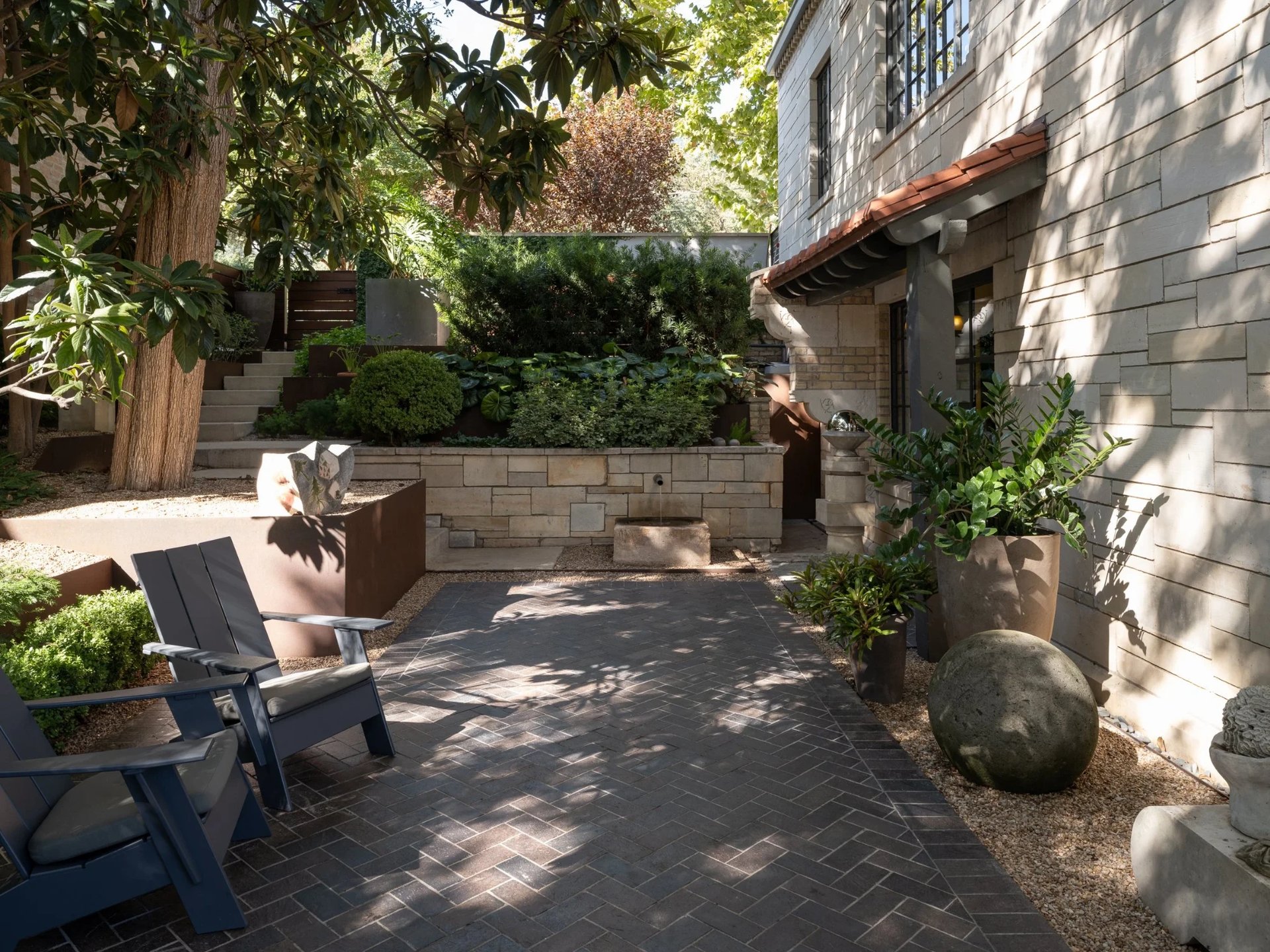 Stone-walled courtyard with blue chairs, a brick path and potted plantings beside a limestone home