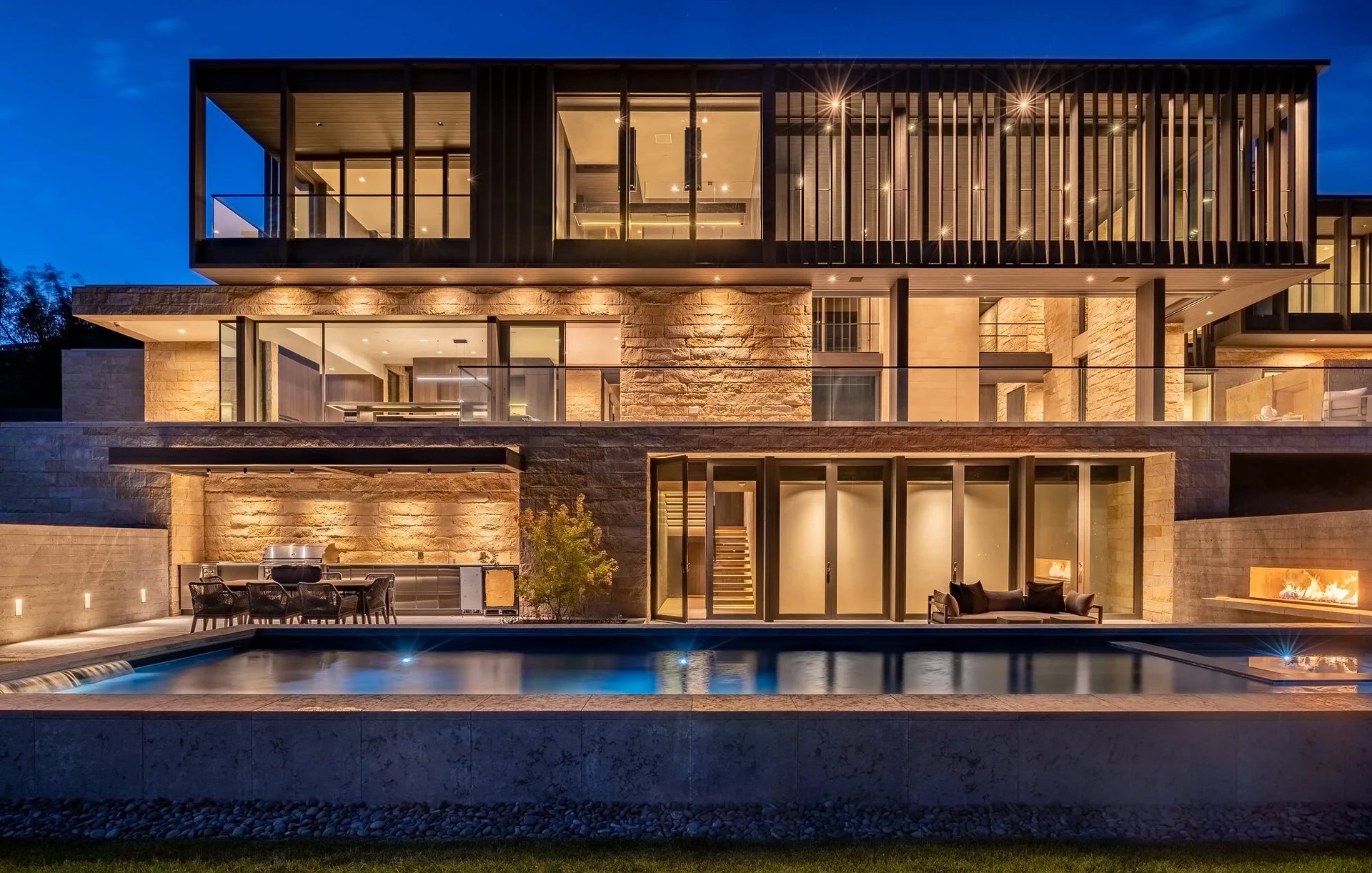 Modern stone home glowing at twilight with a geometric pool, outdoor kitchen, and fireplace in the yard