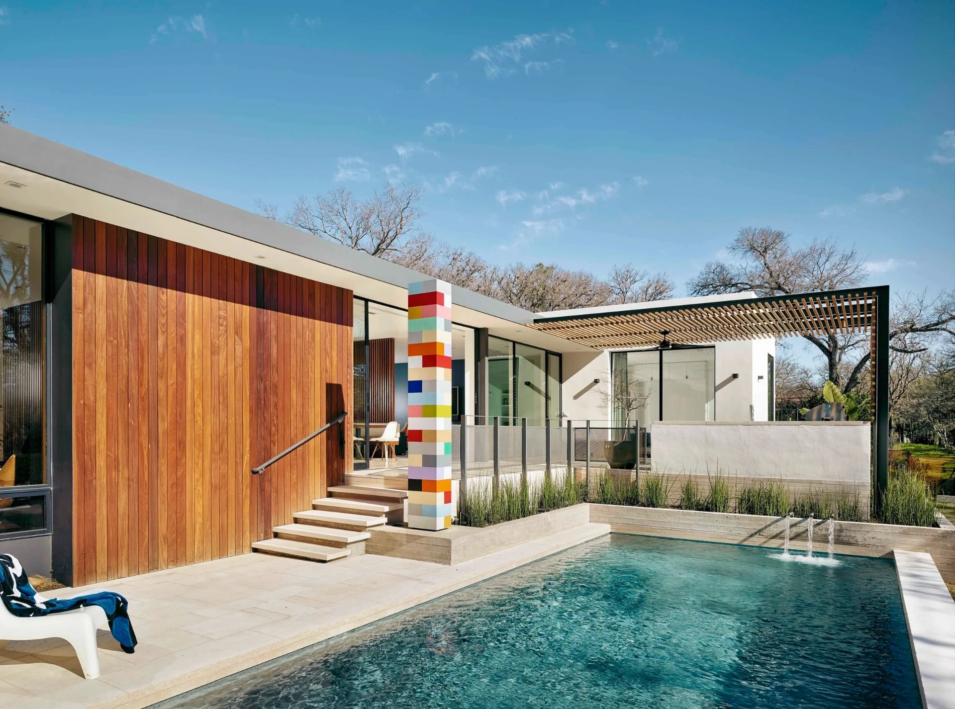Geometric pool beside a modern home with wood siding, pergola, and a colorful tile sculpture