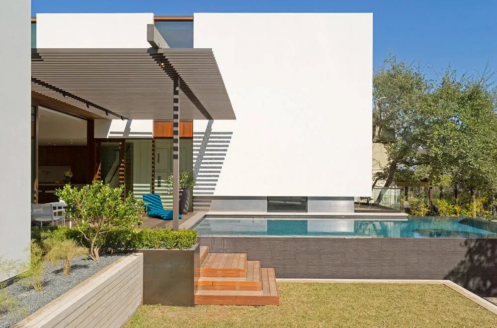 White cubic modern home beside a dark infinity pool with wood steps and a planted retaining wall