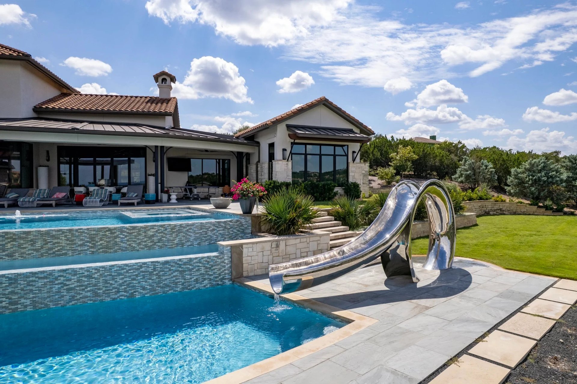 Multi-level infinity pool behind a large Mediterranean home, stainless waterslide on a stone deck