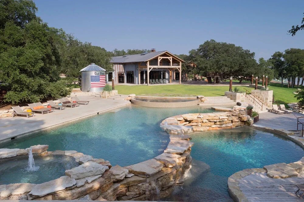 Sprawling freeform pool with rock waterfalls and a wood deck below a Hill Country lodge
