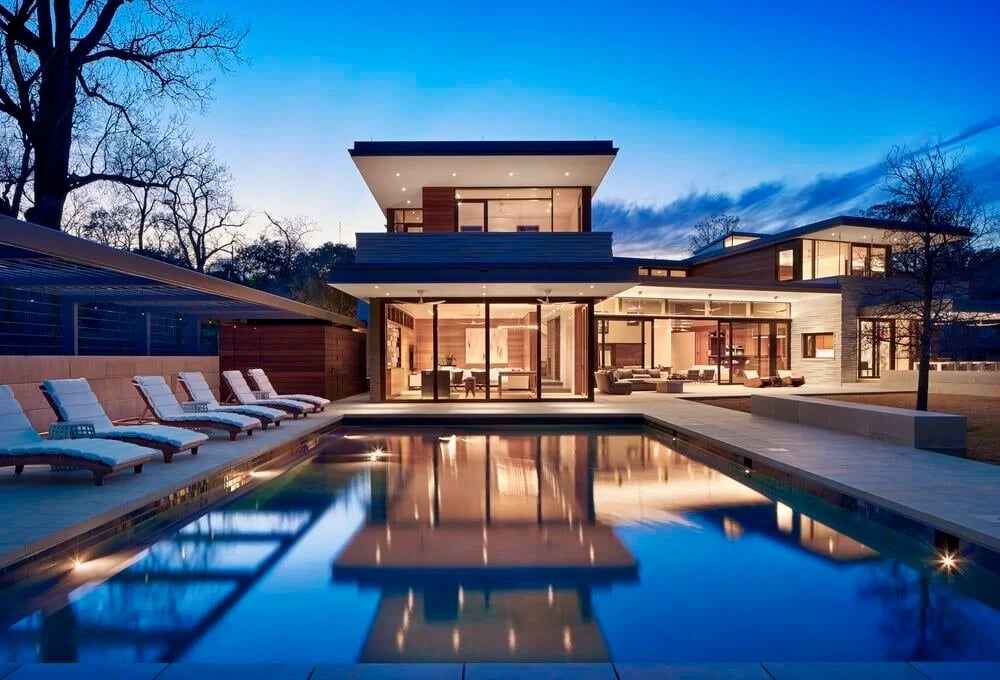 Long geometric pool reflecting a glowing modern home with deep overhangs at blue hour