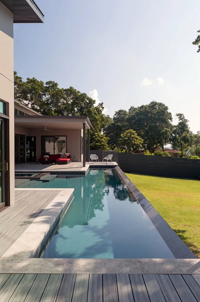 Long infinity pool beside a stucco home, reflecting the sky next to a wood deck and green lawn under tall oaks