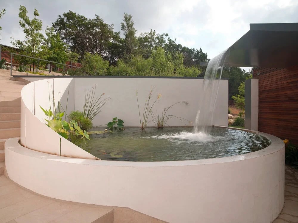 Round raised pond pool with a sheer waterfall, aquatic plants and a curved white wall, limestone steps