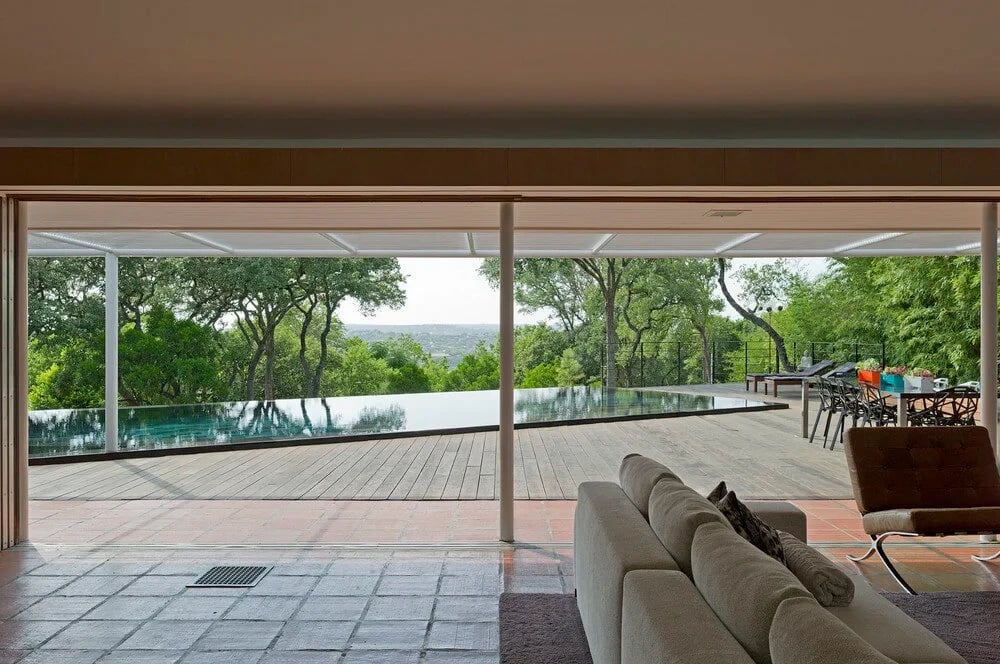 View from a living room through sliding glass to an infinity pool, wood deck and treetops beyond