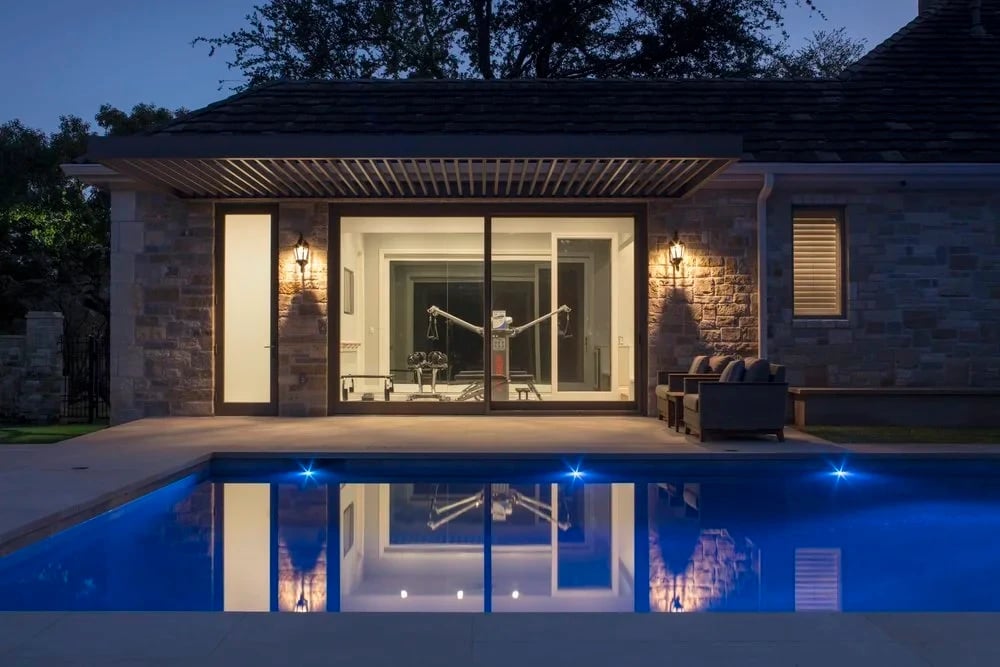 Stone pool house with a pergola and lit sliding doors mirrored in a blue geometric pool at twilight