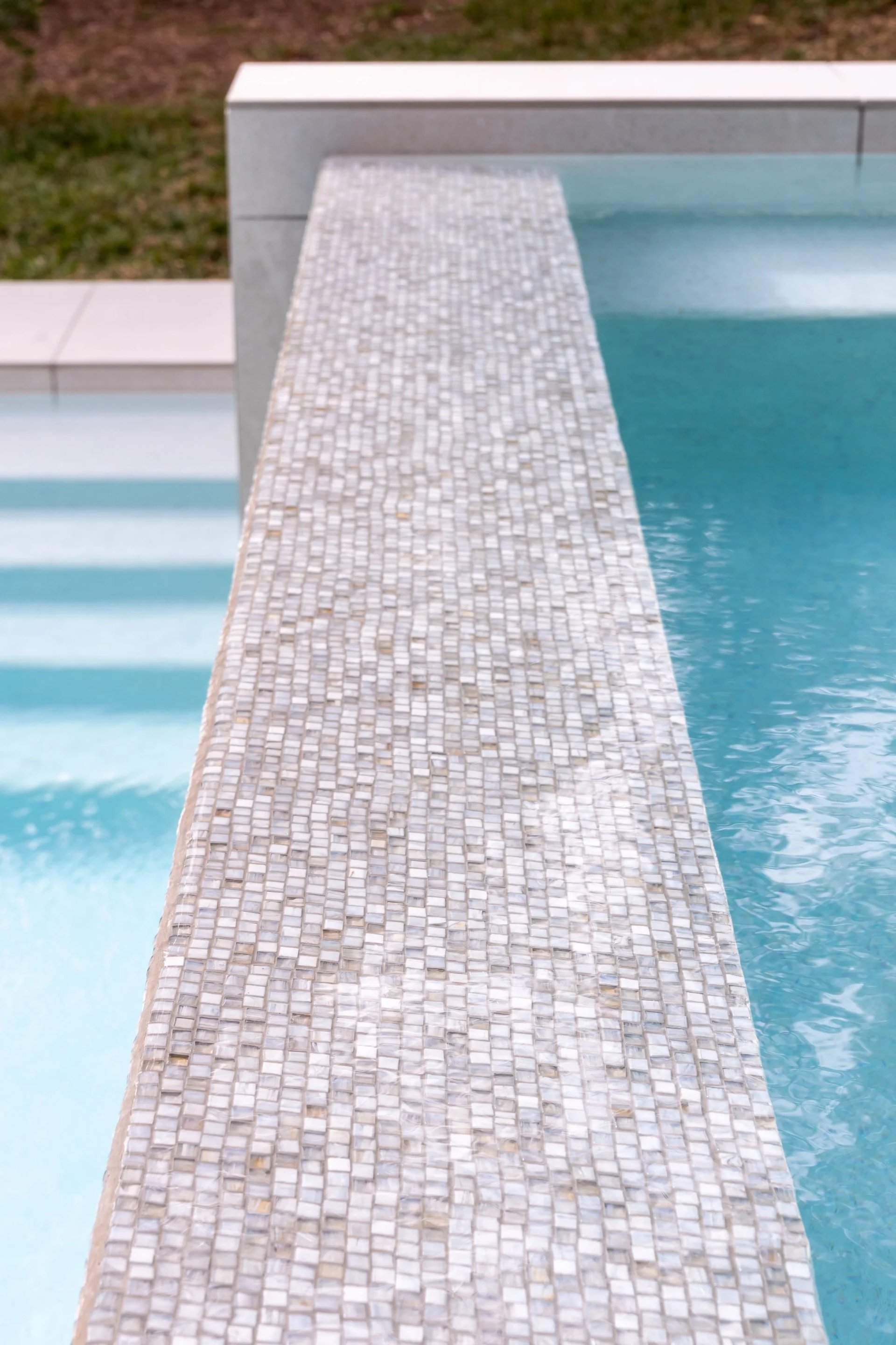 Stone mosaic spillway wall dividing a raised spa from the pool below, with crystal-clear water