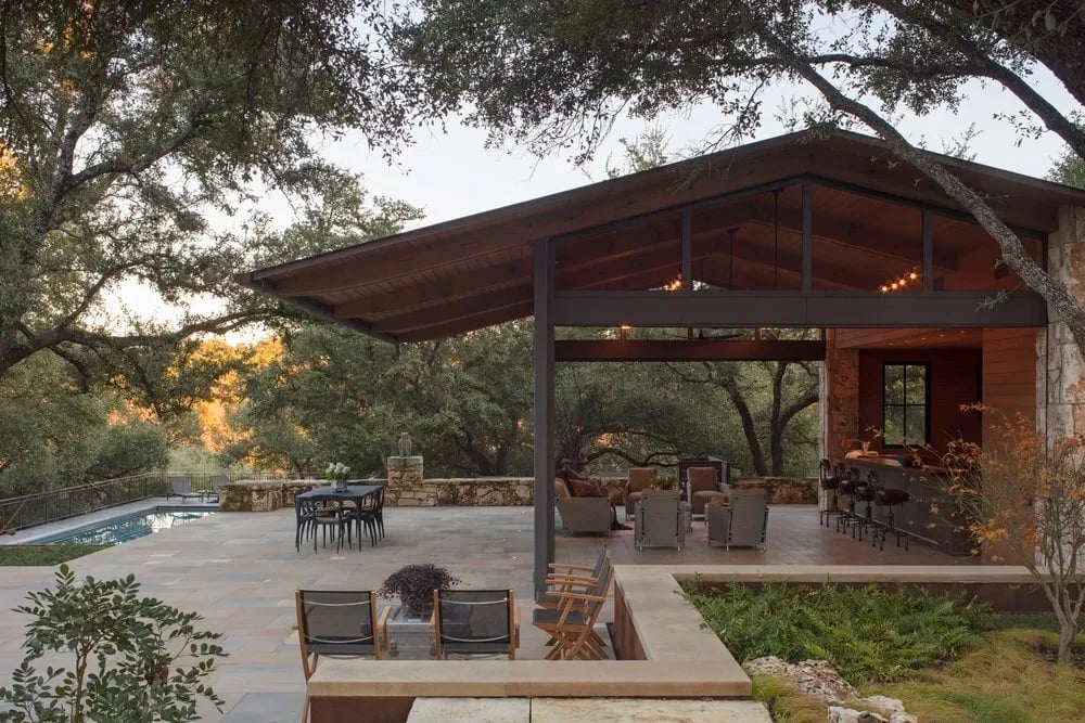 Covered patio with a steel roof, fire pit and seating beside a pool under oaks at dusk