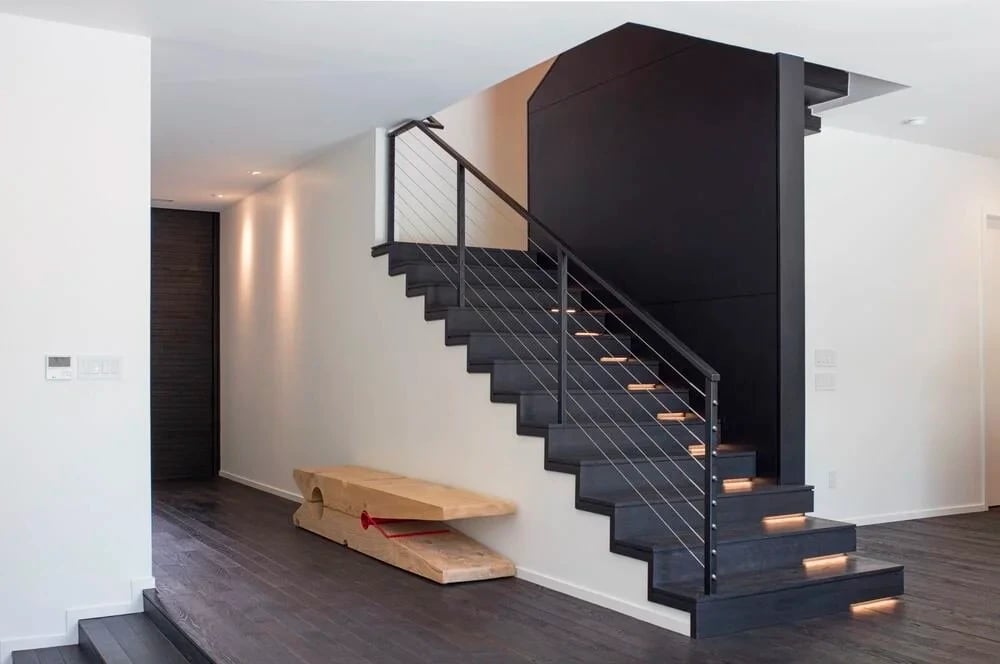 Floating black staircase with cable railing and lit treads against white walls and dark wood floors
