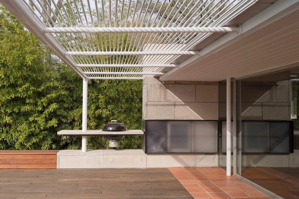 Covered patio with a white pergola, concrete counter and a wood deck against a green hedge
