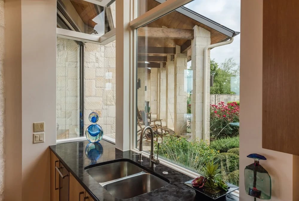 Kitchen sink with a dark stone counter and glass art, framed by a window onto a stone patio and garden
