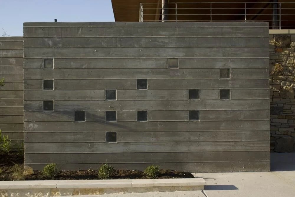 Board-formed concrete wall inset with square glass blocks, beside a stone wall under a timber roof
