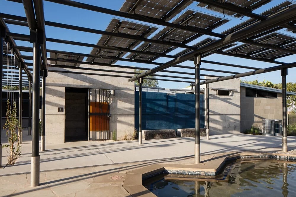 Geometric public pool with a concrete bathhouse and steel pergolas under a clear blue sky