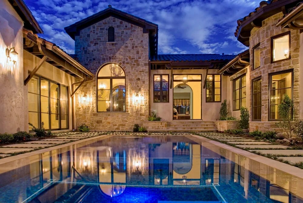 Stone Mediterranean home around a geometric courtyard pool, paver-and-grass deck glowing at twilight