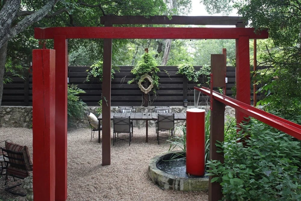 Red steel pergola over a gravel patio with a dining set and circular fountain in a tree-shaded yard