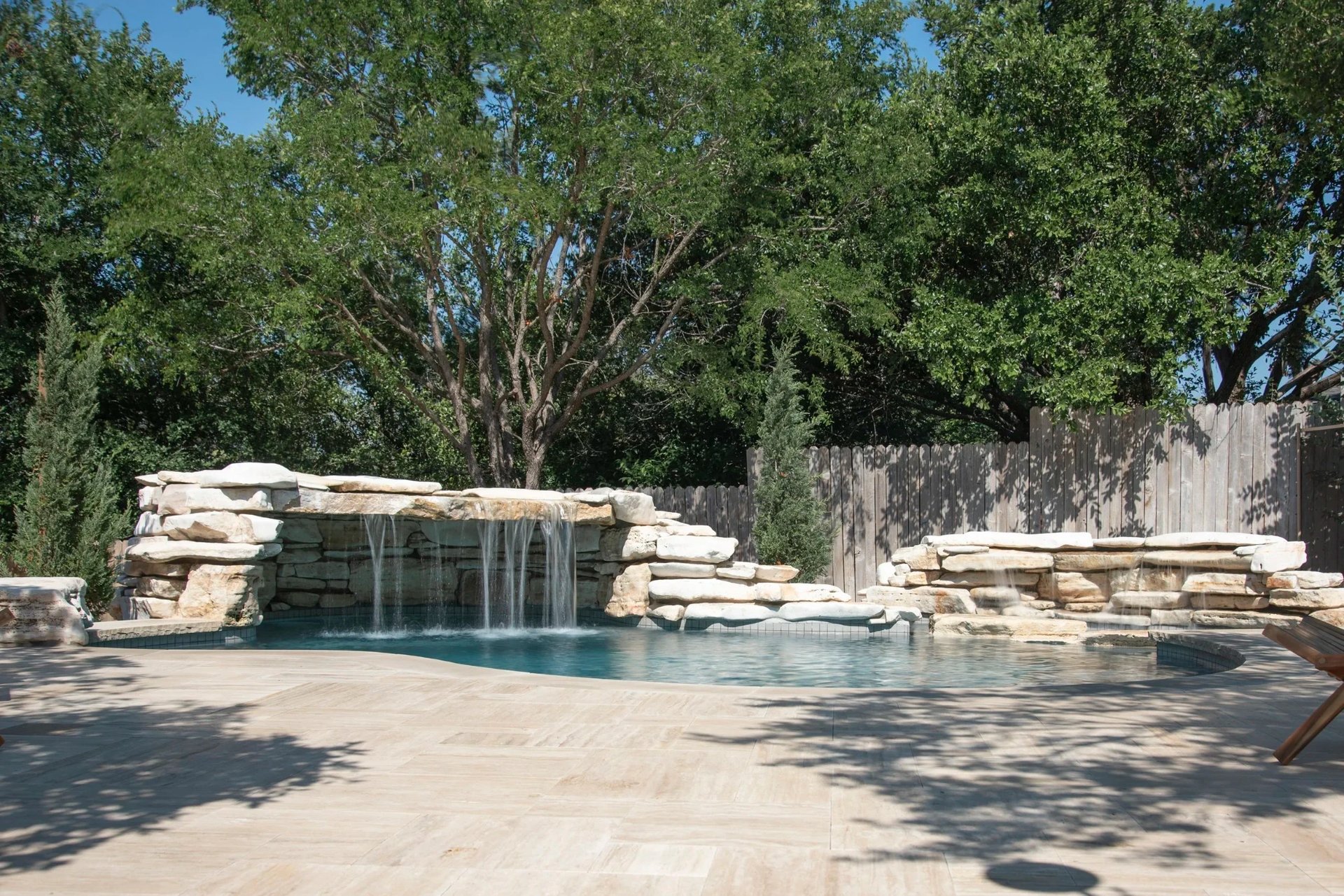Freeform pool with a stacked-limestone waterfall and raised spa framed by trees and a wood fence