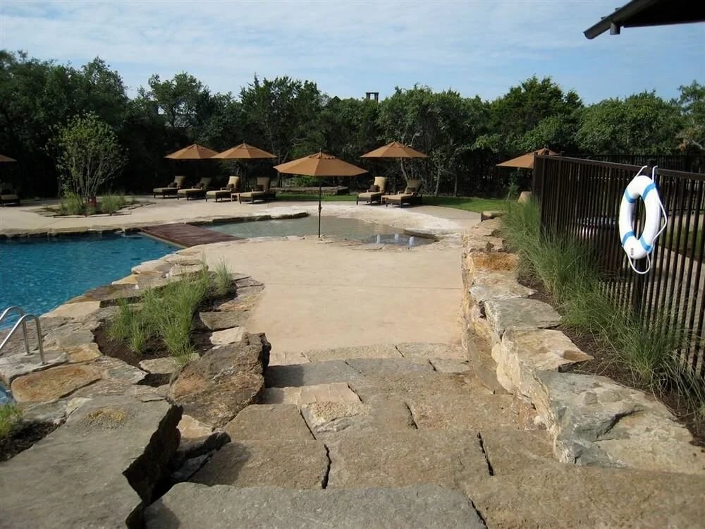 Freeform pool with a flagstone deck, boulders, and umbrellas amid trees in Spanish Oaks