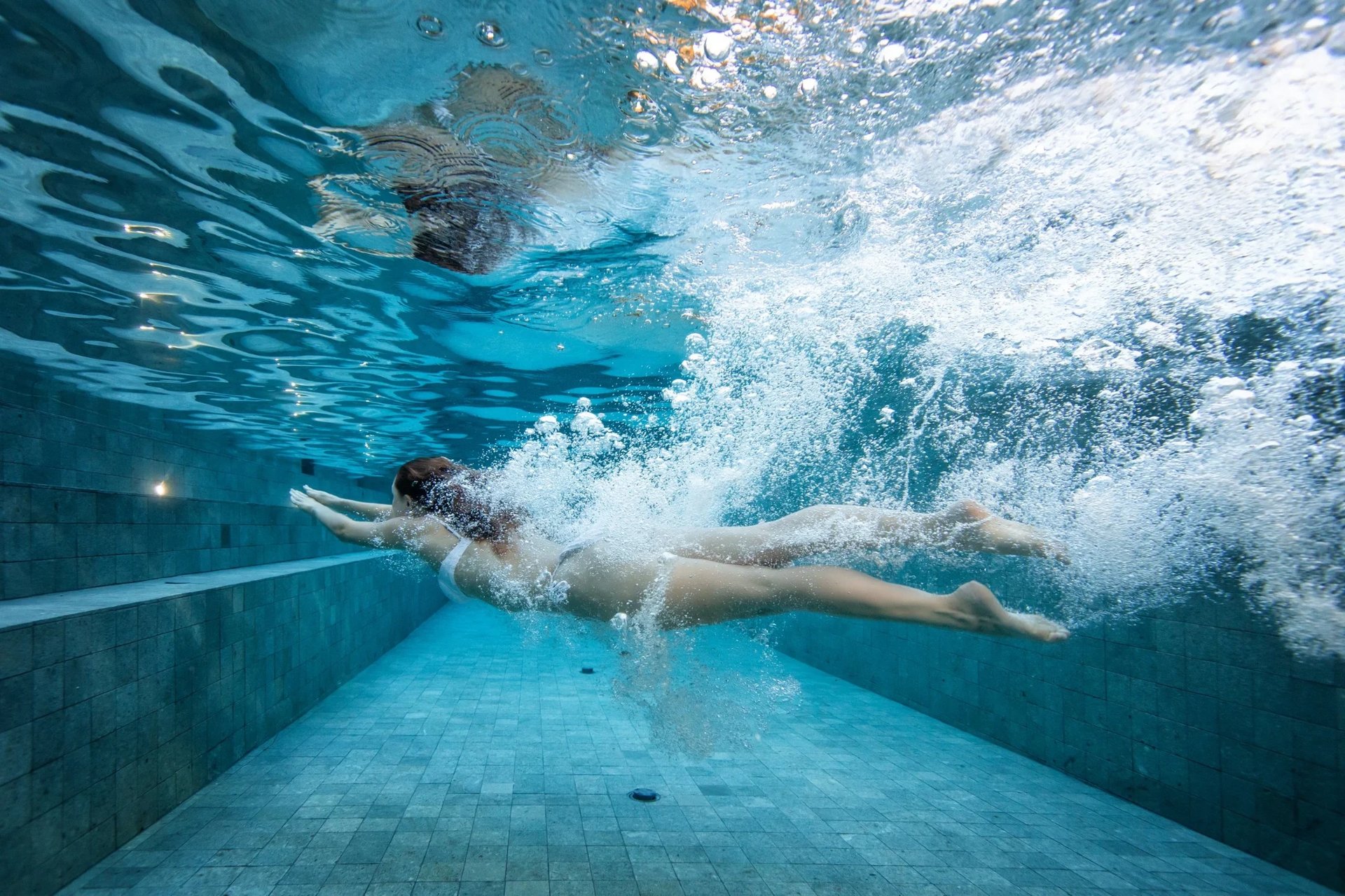 Underwater shot of a woman in white gliding through a tiled pool toward the steps