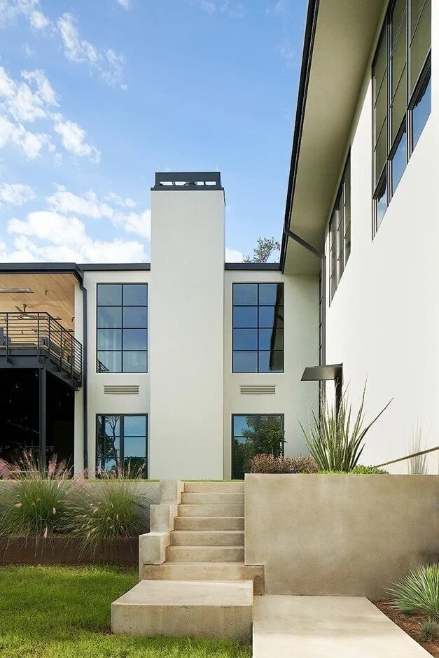 White stucco modern home with a tower and floor-to-ceiling windows above concrete steps and grasses