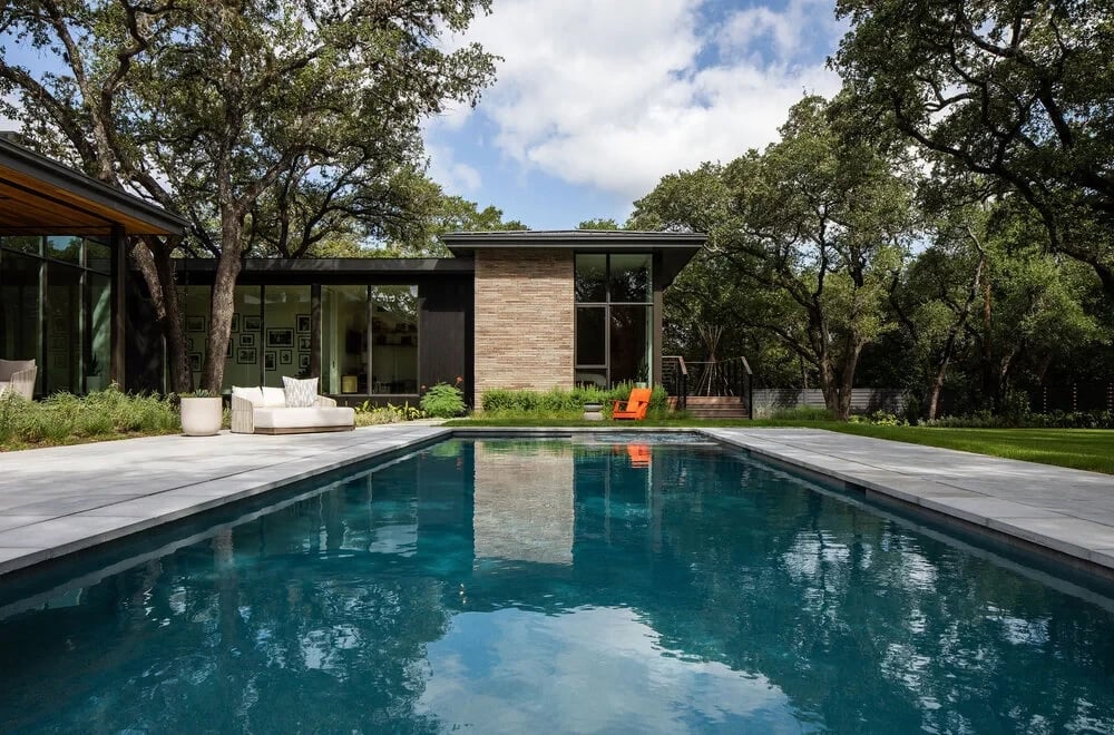 Single-story modern home with walls of glass beside a rectangular pool and lawn under live oaks