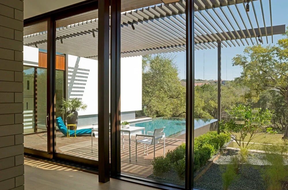 Infinity pool and wooded hillside seen through sliding glass doors from inside a modern home